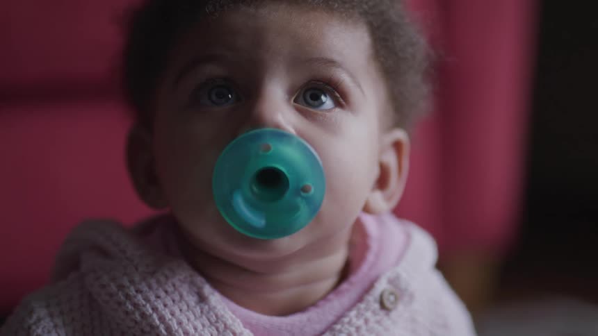 Baby wearing pink and sucking on blue pacifier looks around