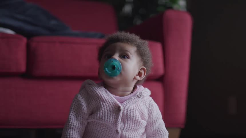 A baby in a pink sweater sits in front of a red couch with a green pacifier in her mouth