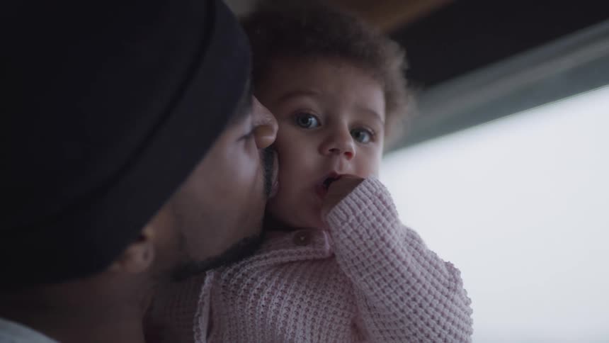 Black father with a beard and a black beanie holds her baby daughter and kisses her cheek