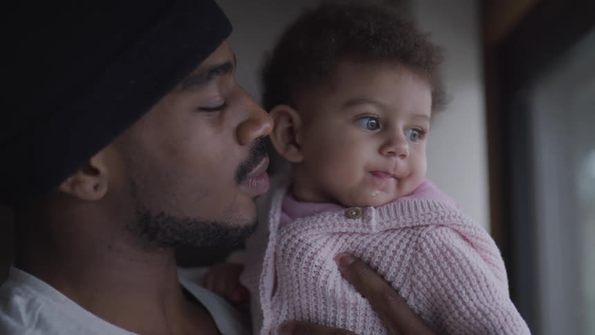 Man wearing knit hat holds young daughter wearing pink sweater as they stand near window