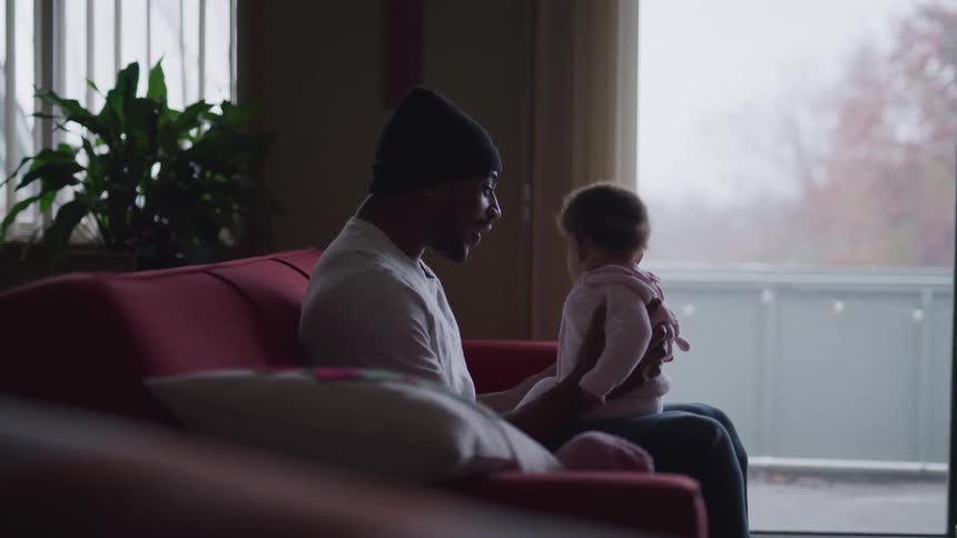 Black man with a beanie and a white sweatshirt sits on a red sofa and holds his toddler girl in the living room at home