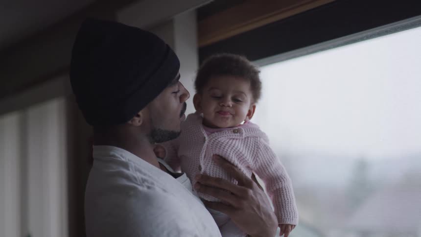 Black father with a black beanie a beard and a white shirt holds his baby daughter with affection