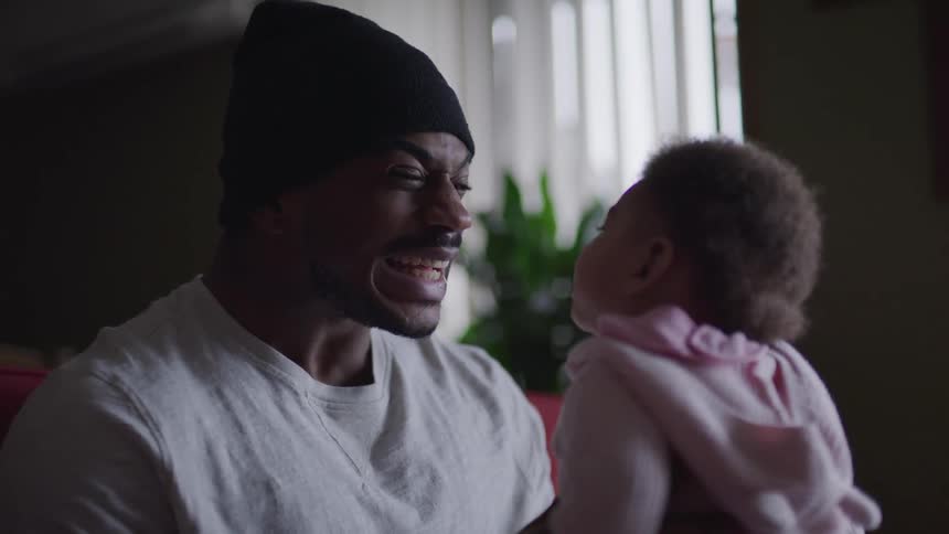 A man with a hat holding a baby and smiling with a plant and a window in the background