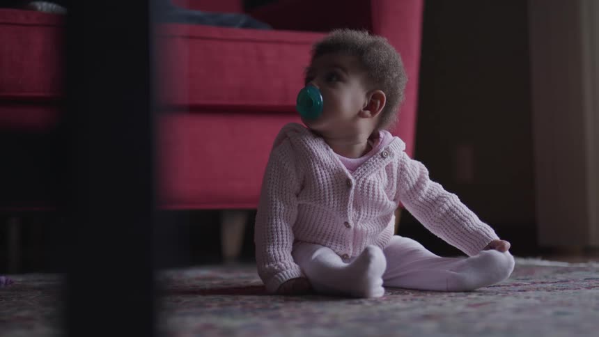Baby sitting on floor alone sucks on their pacifier and holds onto one toe whiling looking up and staring