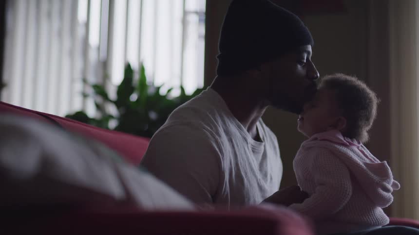 Father with a beanie and a white t shirt holds and kisses his toddler daughter in a room
