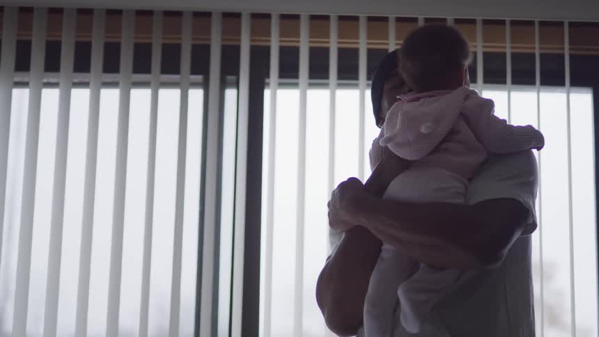 Black father with a beanie and a white t shirt holds her baby in a room