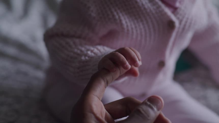 A baby sitting on a bed grips the finger of an adults hand