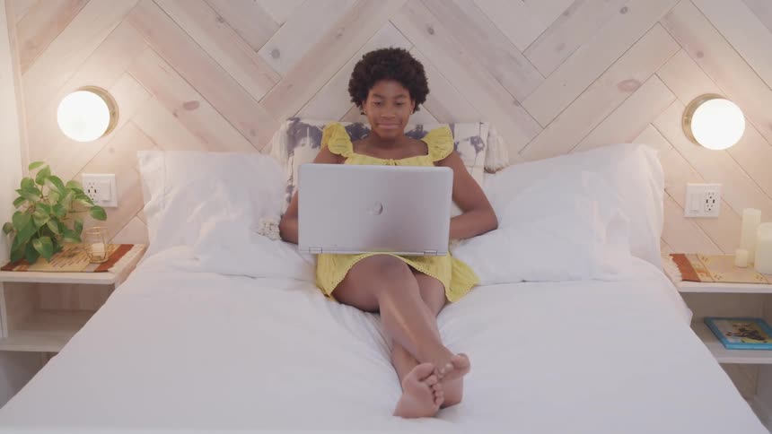 Black woman with curly hair and a yellow dress sits on a bed and uses a grey laptop in the bedroom