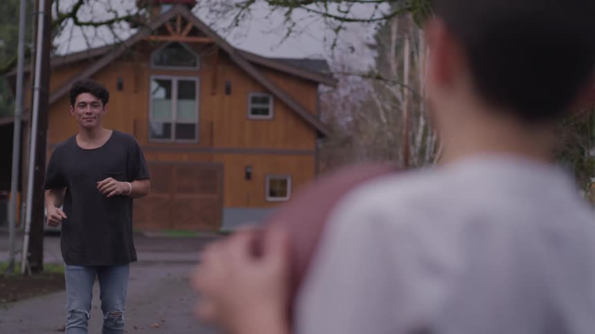 A boy holds a football standing in a driveway and a teenager brother anticipates a throw