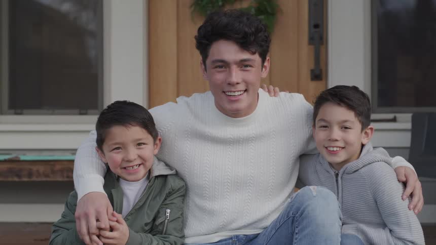 A teenager sits on the front porch and has his arms around two younger brothers posing for a photo