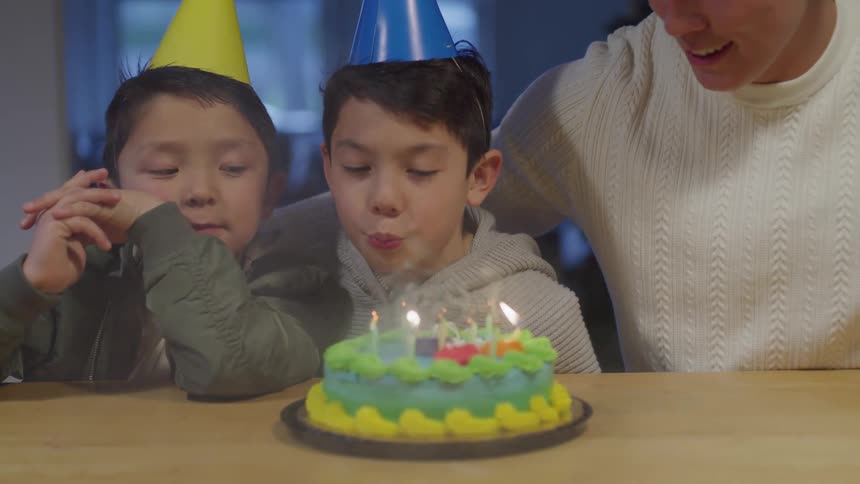 Two young boys sit with their father as one blows out the candles on a birthday cake