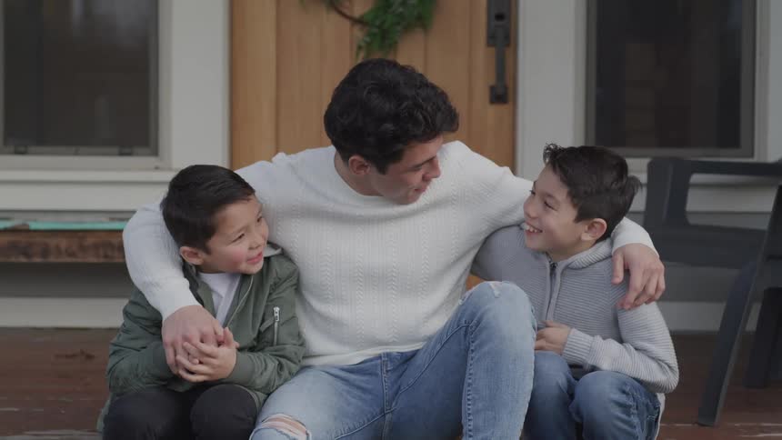 A teenage boy sits on the front step of a house with his arms around his little brothers and they talk and laugh