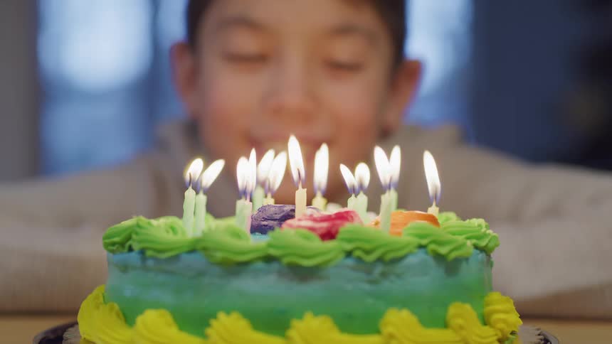 A boy rests with his elbows on a table as he watches the candles burning atop a colorful cake
