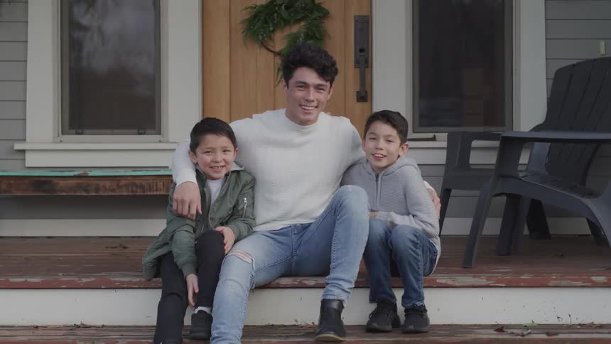 A teenage boy sits on the front step of a home posing for a photo with younger brothers