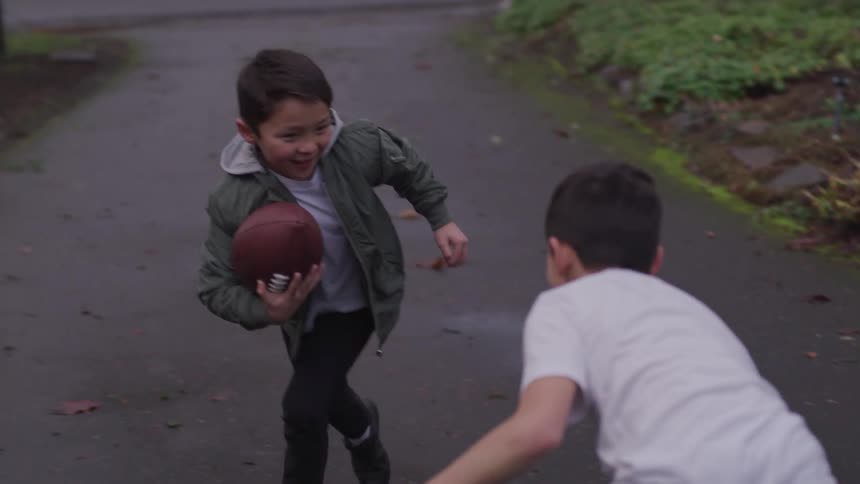 A young boy runs with a football and smiles as he tries to avoid his brother and father