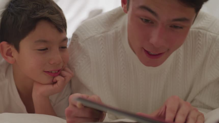 A man and a boy laying on a bed looking at a tablet
