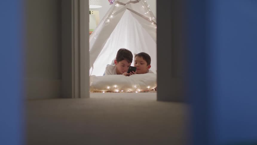 Two young boys lie in a fort in their house and play with a cell phone