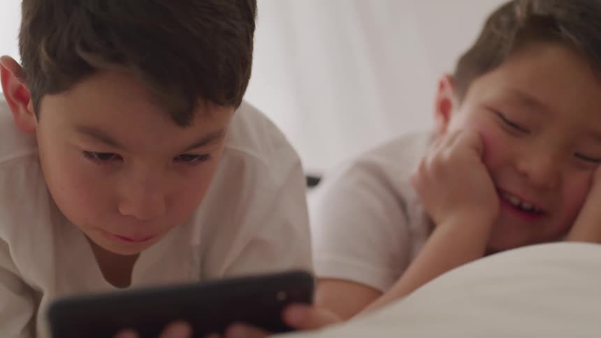 A pair of boys lie on their stomachs on a bed as they look at a tablet