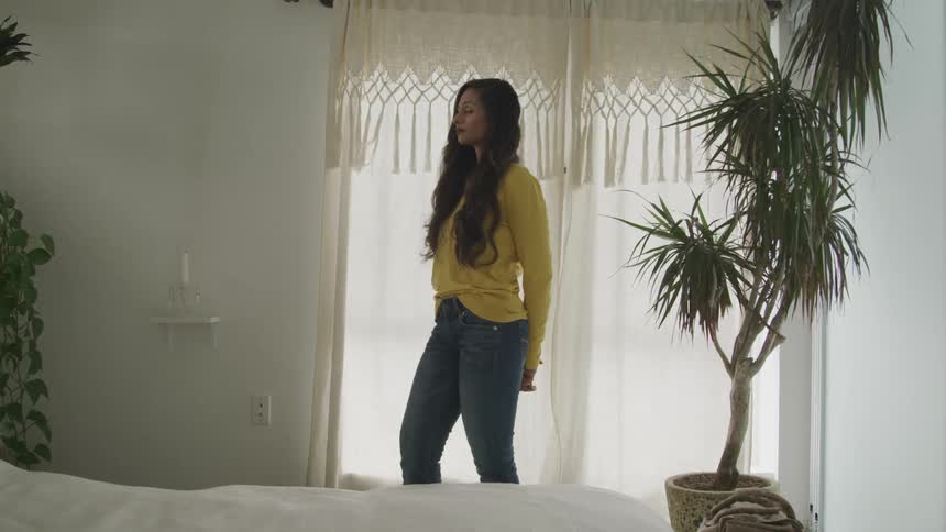 A woman with long hair stands in a bedroom near a potted plant