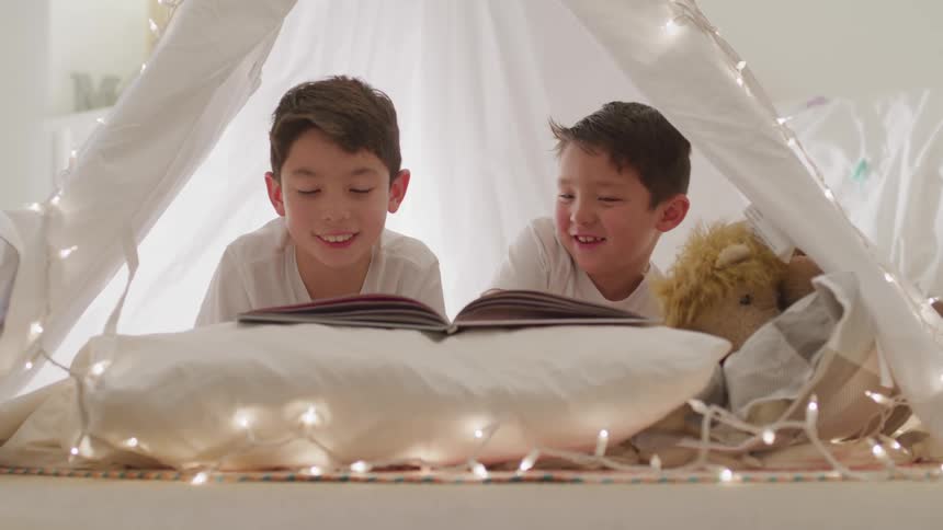 Two male siblings are enjoying reading a book together in their makeshift bedroom fort