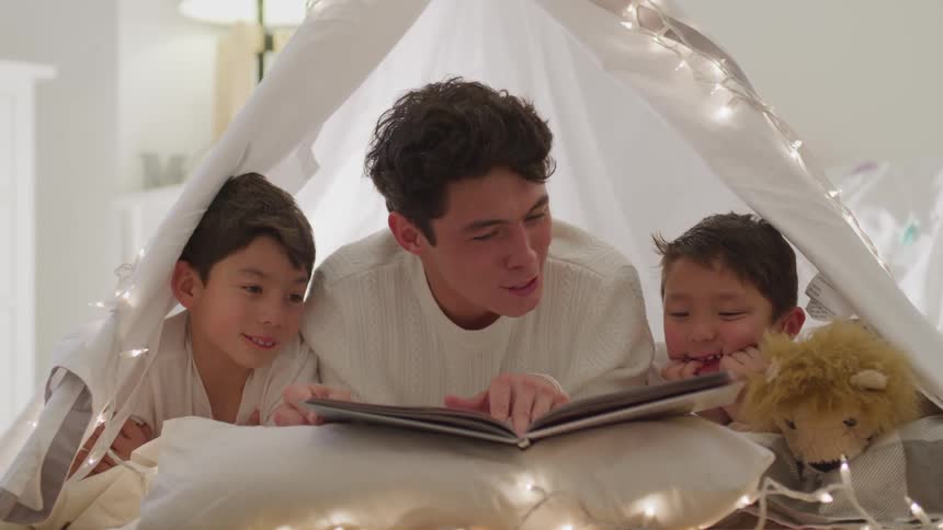 A teenage boy lays inside a homemade tent from a sheet and reads a book to younger brothers