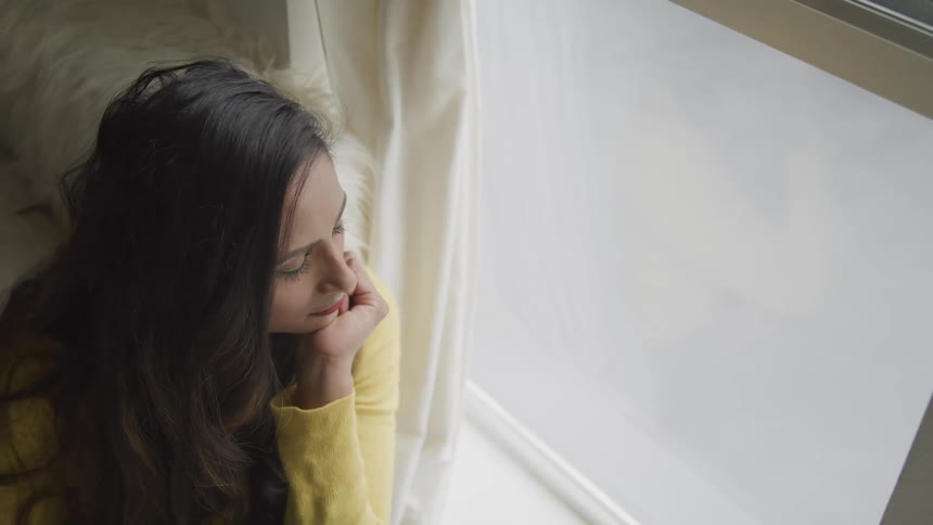 Brunette woman with black wavy hair and a yellow sweater looks out the window