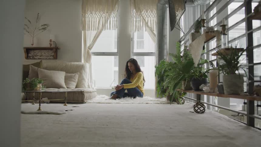 A woman sits silently curled into a ball in a corner window area of a her living room