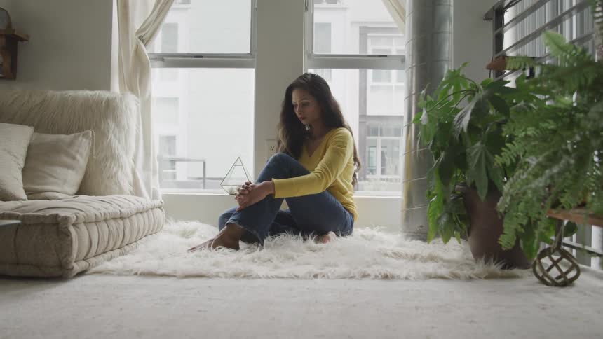 Woman sits on fuzzy white rug next to living room window showing opposite building