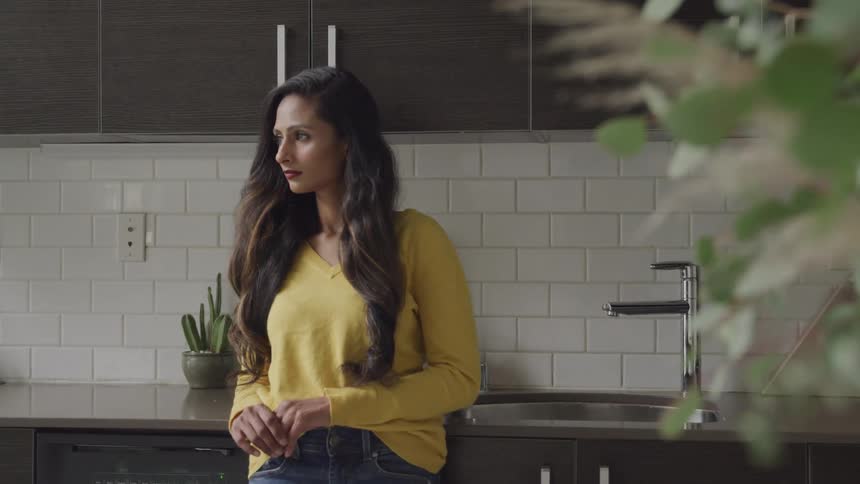 Brunette woman with wavy long hair a yellow sweater and jeans leans against kitchen counter in the kitchen