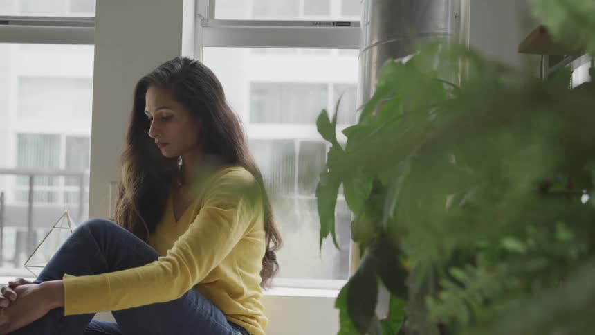 A woman with a sweater and jeans sitting next to a window with plants in the foreground