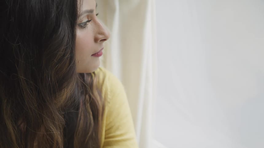 Long haired brunette woman briefly leans with chin on hand while looking out curtained window