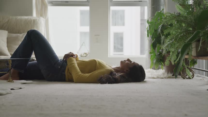 Brunette woman with jeans and a yellow sweater lies on a carpet in the living room