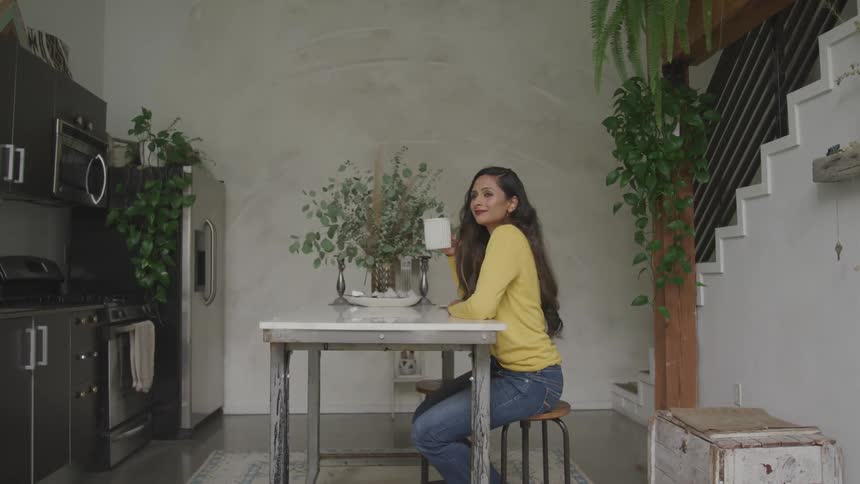 A woman sits on a stool at a dining table in a small kitchen