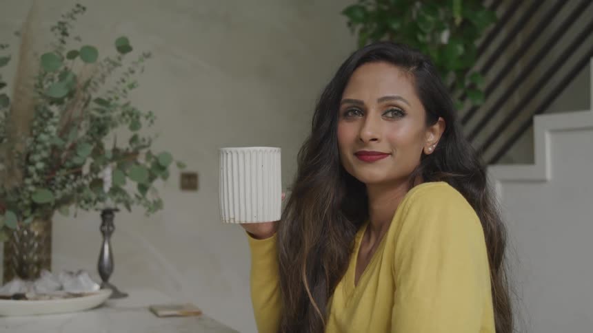A woman with long hair sits at a table while holding a large coffee mug
