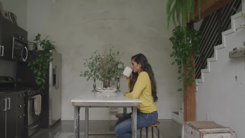 Brunette woman with a yellow sweater jeans sits at a table and drinks a cup of coffee in the kitchen