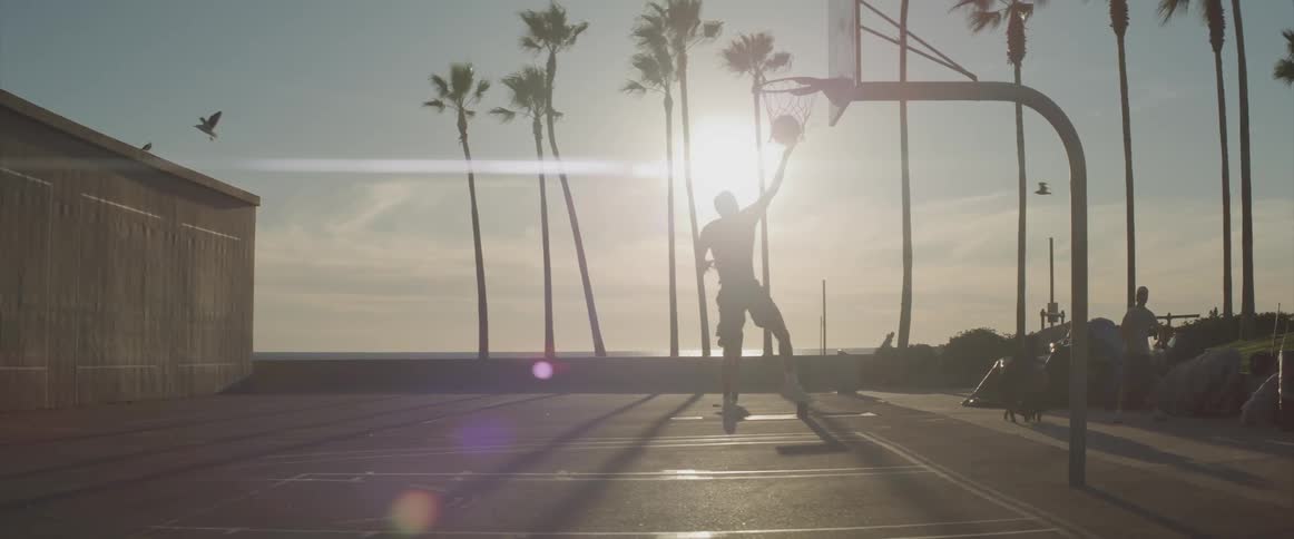 A man on a basketball court with palm trees around it on a sunny day is doing a layup with several people on watching him