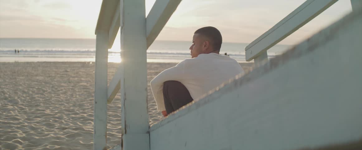 Young man sits on wooden ramp on beach in front of setting sun