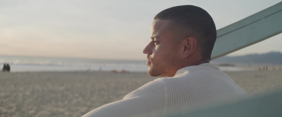 Man sits on beach near white painted rail looking out toward sea
