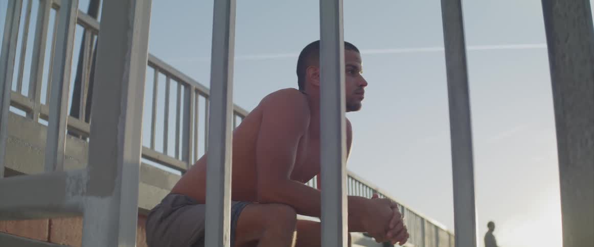 A young man sits on an outdoor seat and looks outward in contemplation under a bright sun