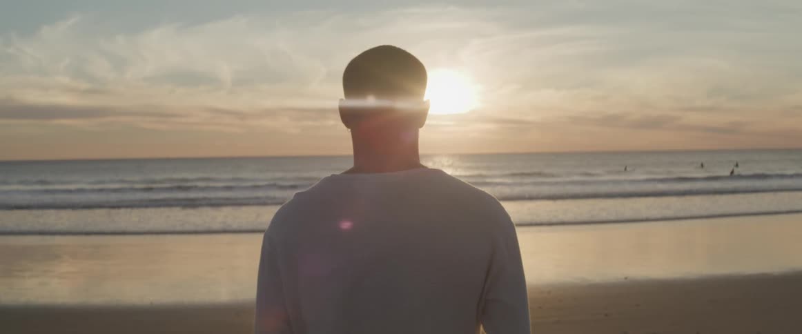 A man stands on the beach and looks at the water as the sun sets
