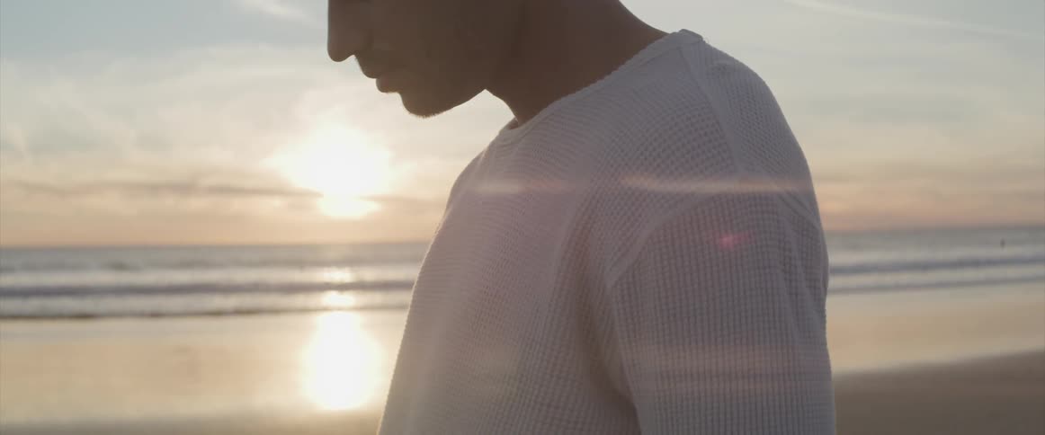 Man with a white knit sweater looks down on the sand beach at sunset next to the sea