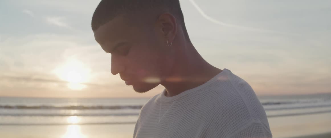 Side view of a man with earring and a white sweatshirt standing on the wet beach at sunset
