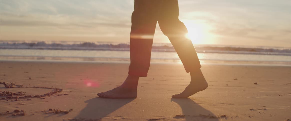 Man with black pants walks barefoot along the beach at sunset