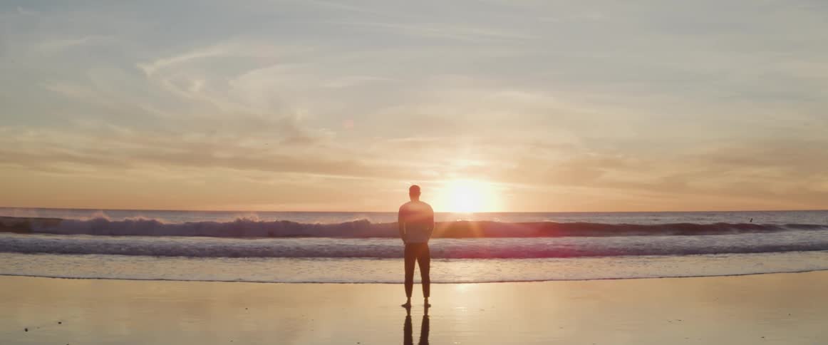 Man standing barefoot on the wet beach is silhouetted against the sunset