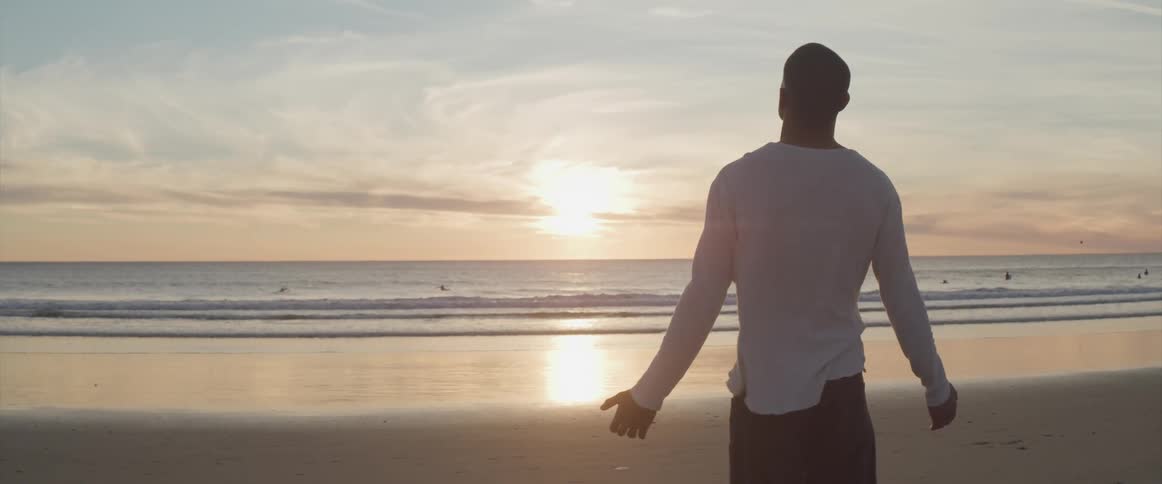 A man stands on the beach at sunrise and raises his arms up and out