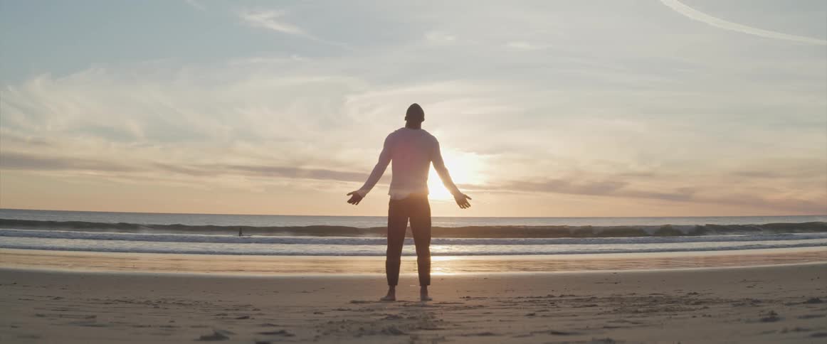 A man stands on the beach and reaches his arms out to his side as the sun rises