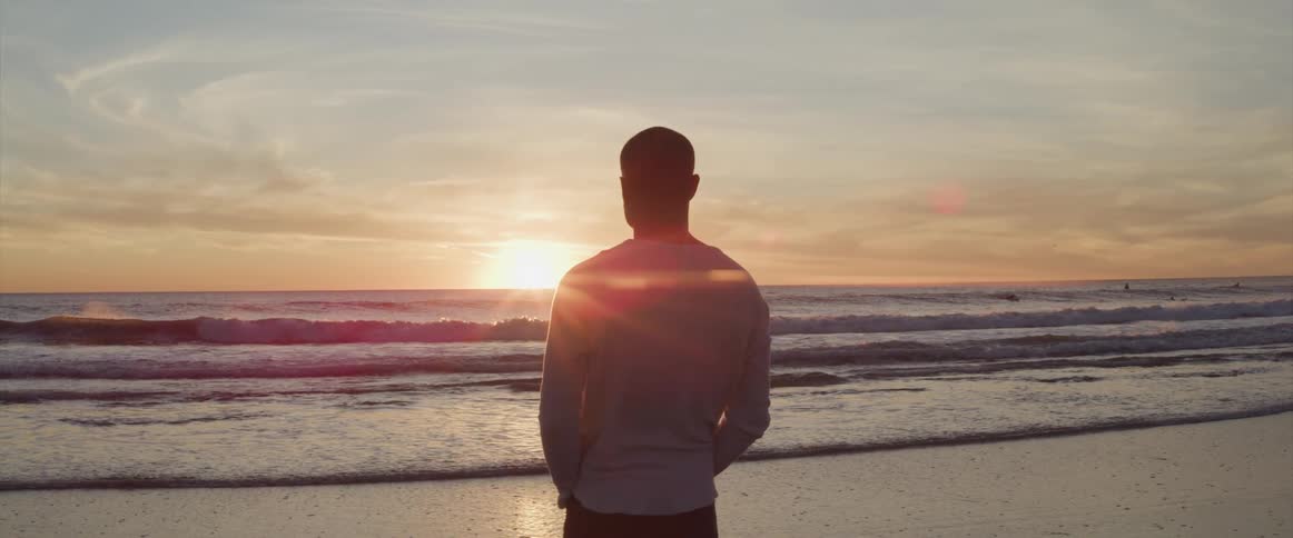 Man standing barefoot on the wet beach is silhouetted against the sunset