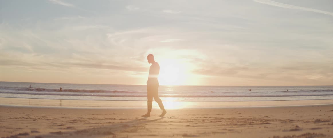 A man walks barefoot along the beach with his hands in his pockets as the light of the low sun cloaks half his body in shadow