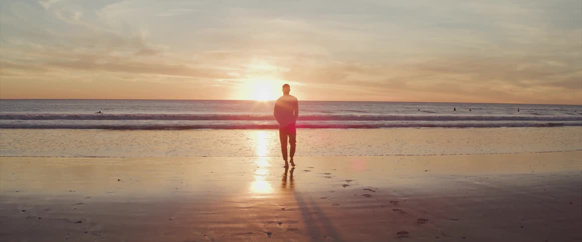 A man walks away from the ocean on the beach as the sun sets behind him