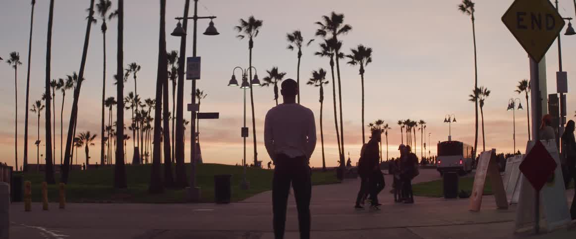 Man stands in busy park with hands in pockets looking toward red sky